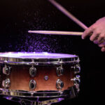 Drum sticks hitting snare drum with splashing water on black background under studio lighting close up.